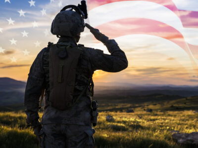 Soldier saluting a transparent US flag while facing the sunset